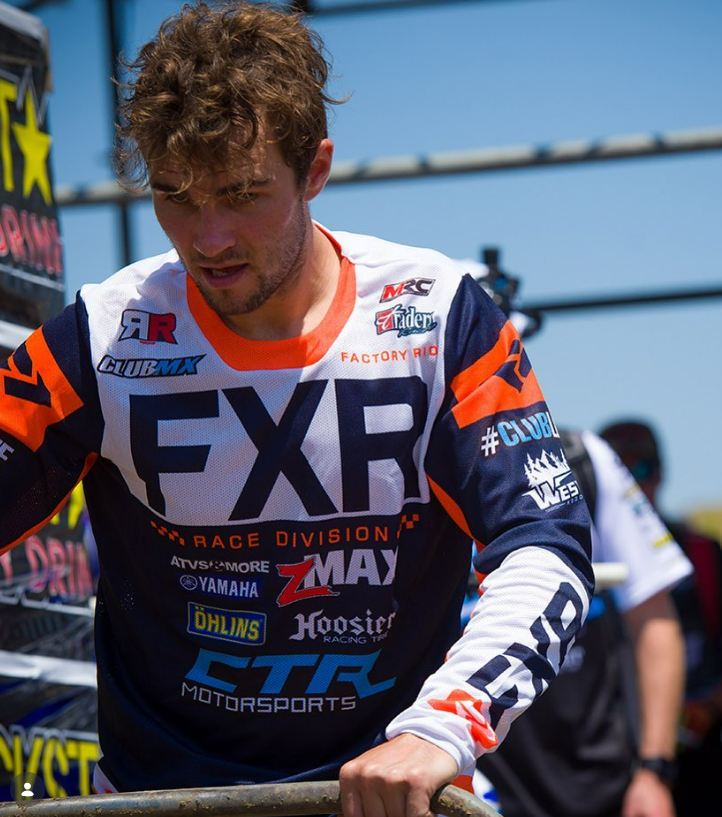 A young male motocross rider with curly hair, wearing a MX racing jersey with various sponsor logos, standing outdoors in front of a race setup