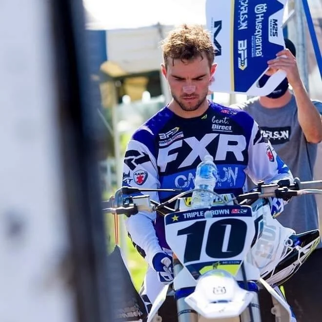 A young man in motocross gear sitting on a dirt bike, with a water bottle on the bike's front.