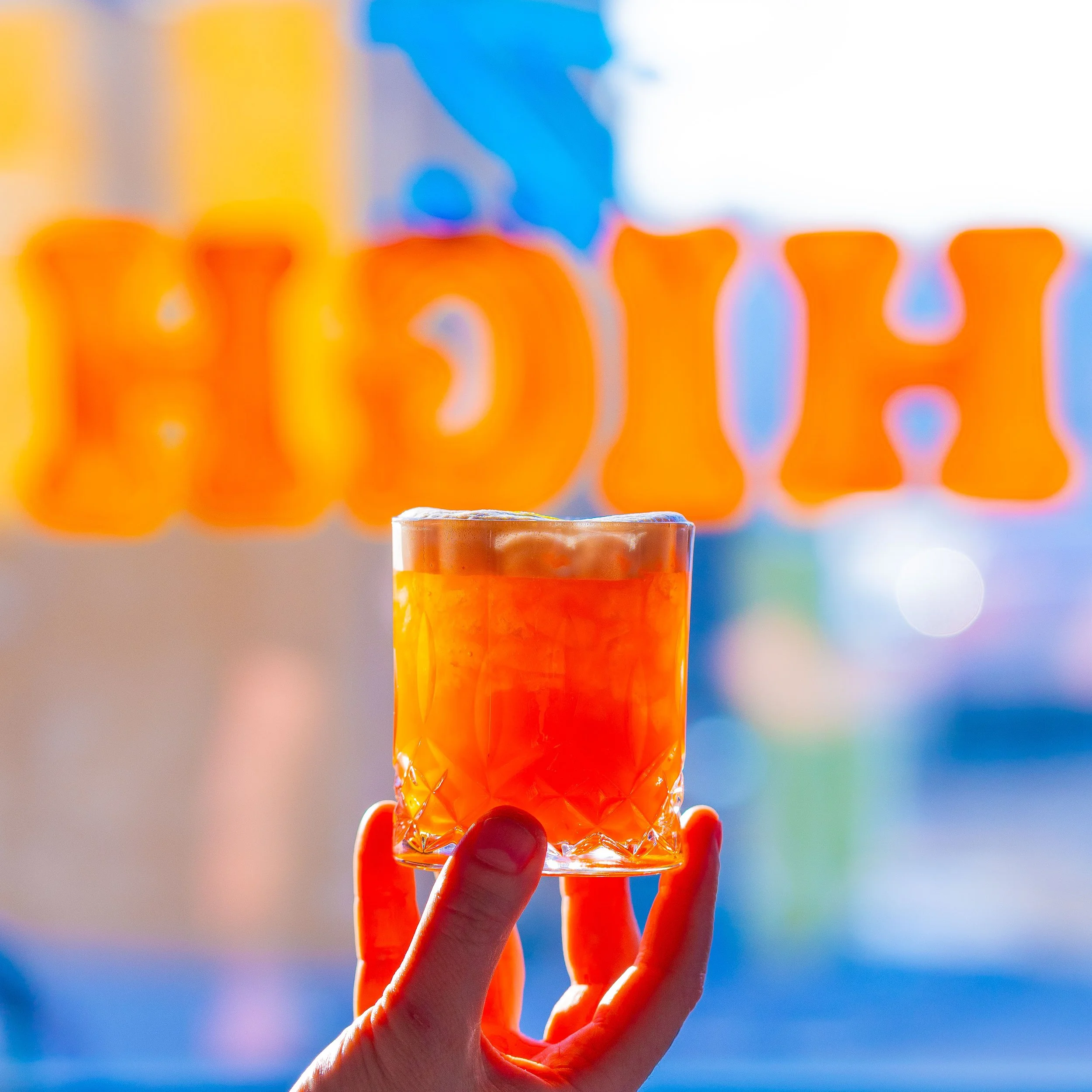 A hand hold an orange cocktail in front of a hand painted window sign