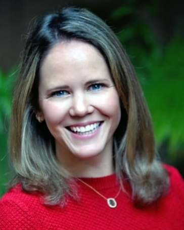 A woman with shoulder-length light brown hair, wearing a red top and a gold necklace with a small pendant, smiling outdoors with green foliage in the background.