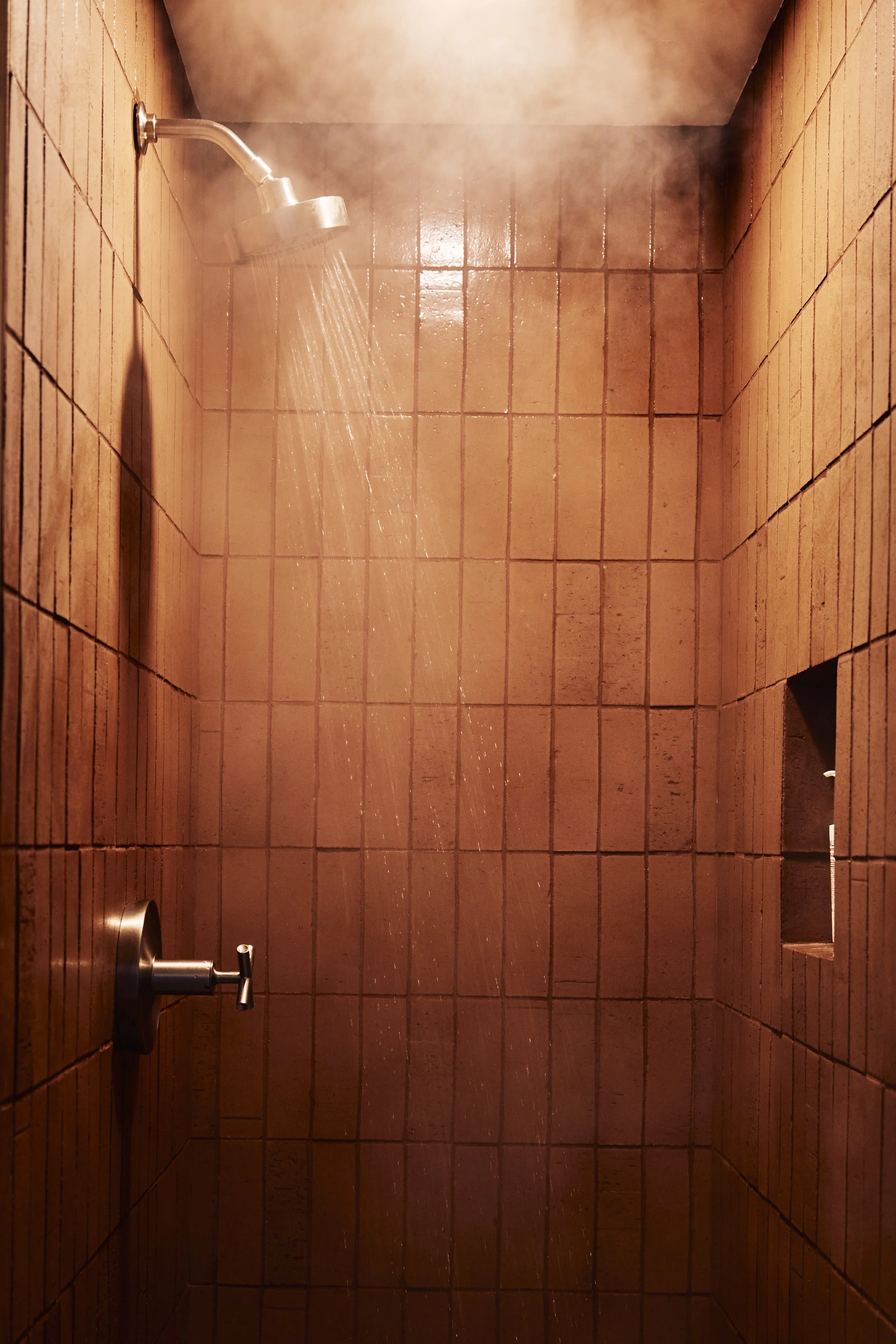 A shower with a metal showerhead and faucet on a tiled wall, with water running and steam rising.