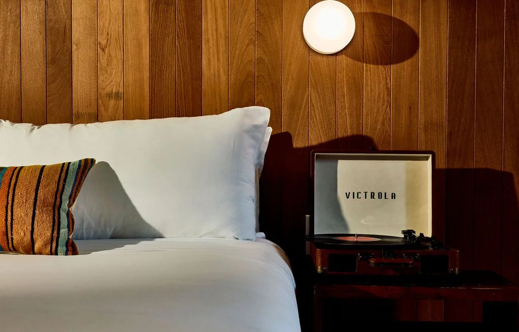 A bedroom with a wooden-paneled wall, a white bed with a colorful striped pillow, and a vintage Victrola record player on a dark wooden nightstand.