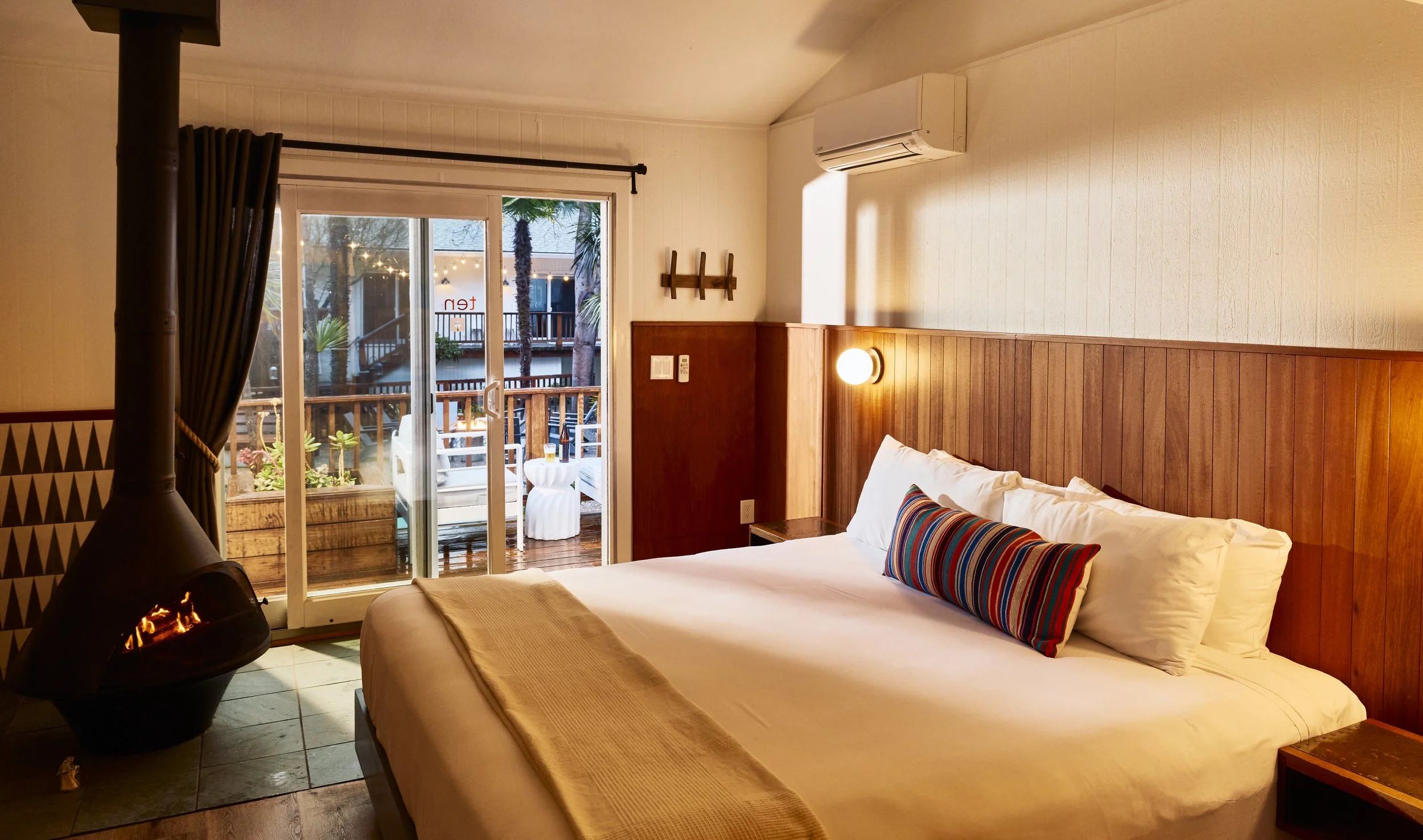 A cozy hotel room with a large bed, colorful striped pillow, wooden wall paneling, and a balcony with outdoor seating, overlooking palm trees.