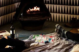 People playing a board game on the floor near a lit fireplace with a bottle of wine nearby.