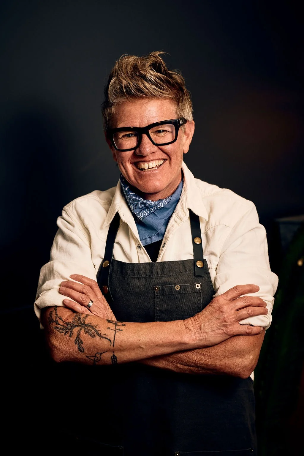 Smiling woman with short hair, black glasses, tattooed arm, wearing a white shirt, black apron, and blue bandana, standing with arms crossed against a dark background.