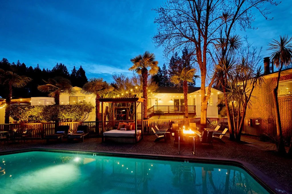 An evening backyard scene with a swimming pool, outdoor fire pit, lounge chairs, palm trees, and a house in the background under a blue sky.