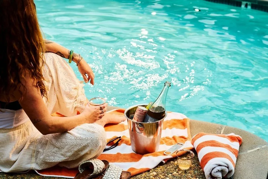 A woman sitting by a poolside with a drink in hand, a striped towel, and a metal bucket with bottled beverages, surrounded by a rolled towel and sunglasses.