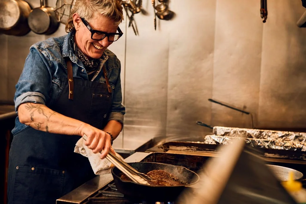 A smiling person cooking on a stove in a kitchen, wearing glasses, a denim shirt, and an apron.