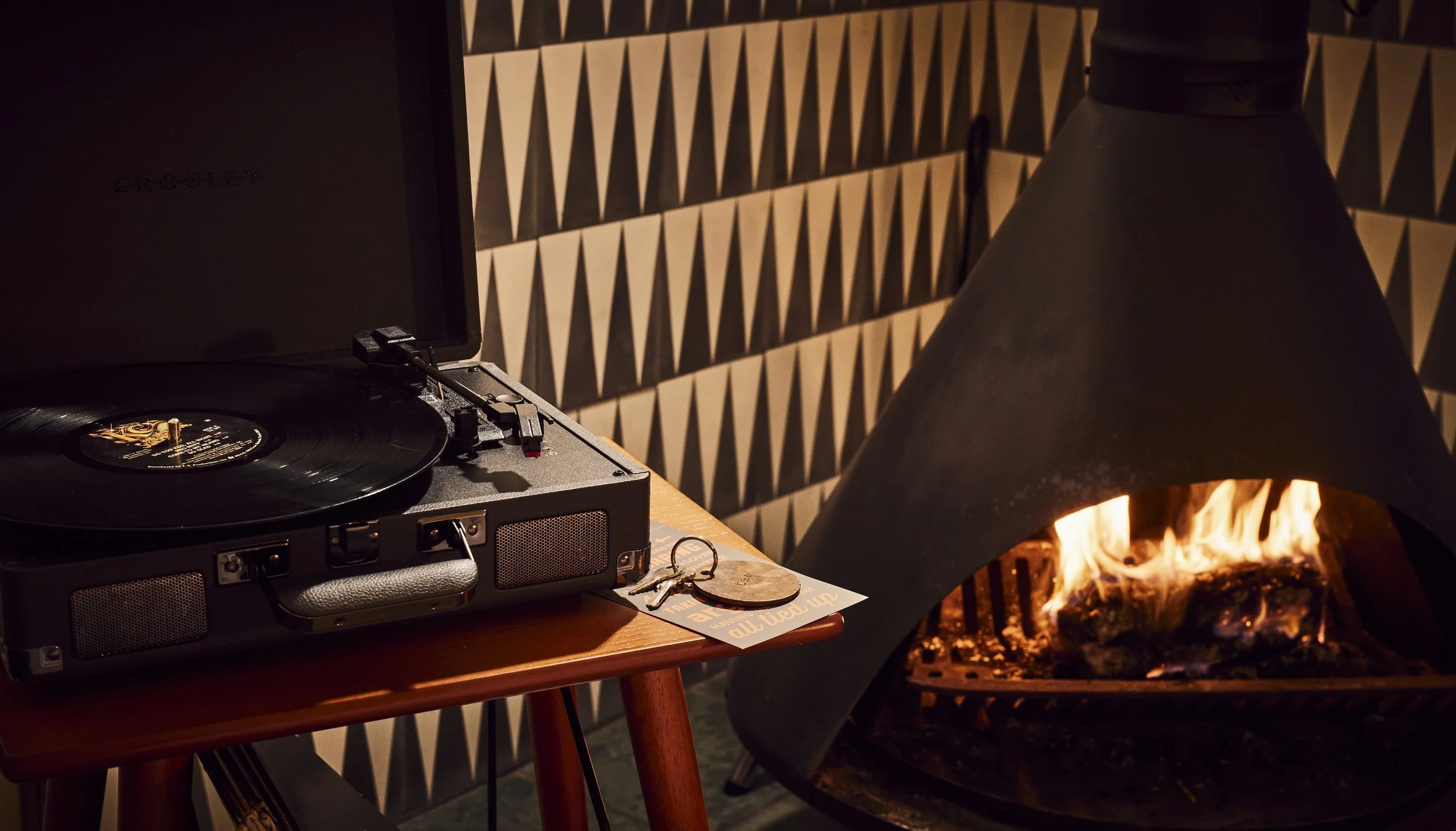 A turntable playing vinyl records on a wooden table, with a pair of keys and a circular object next to it. To the right, there is a lit fireplace with a black metal hood and a geometric patterned wall in the background.