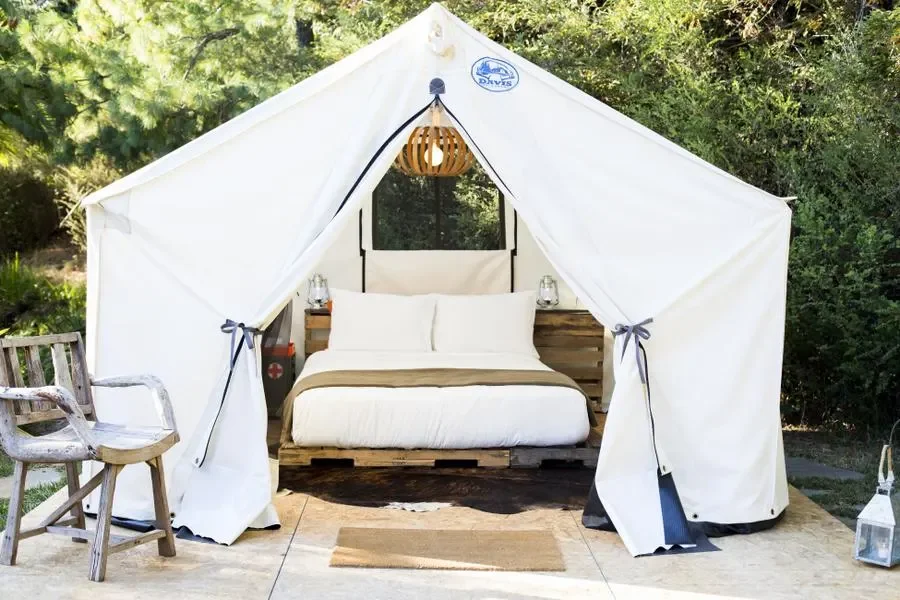 Canvas tent with a bed inside in a nature setting, with a wooden chair, small side tables, and decorative lanterns nearby.
