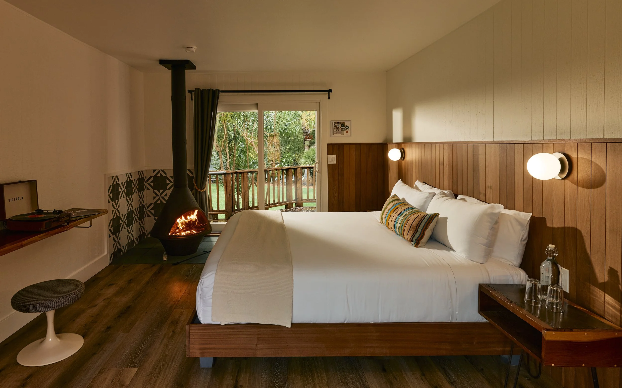 A cozy bedroom featuring a bed with white bedding and a striped pillow, wooden wall accents with mounted lights, a glass door leading to a balcony with greenery outside, a fireplace in the corner, and a small table with glasses next to the bed.