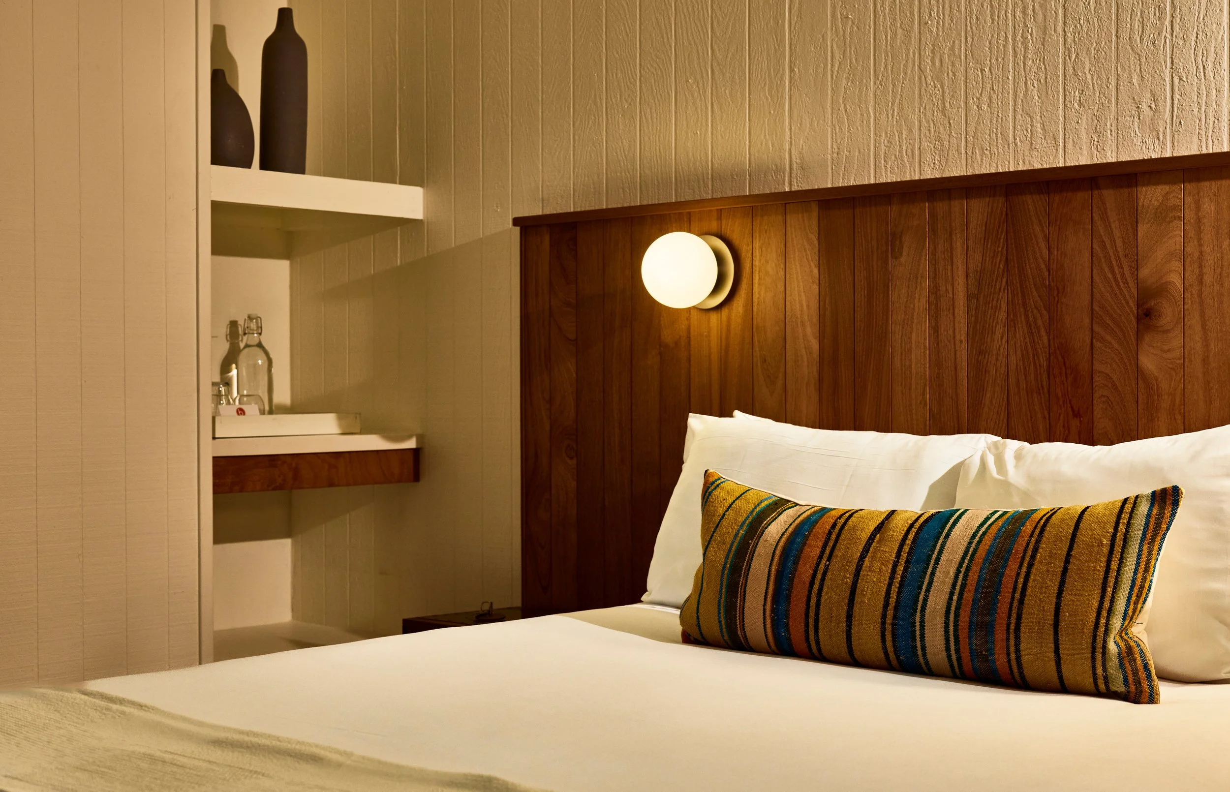A bedroom with a wooden headboard, a white wall-mounted light, a bed with a colorful striped pillow, and a built-in shelf with decorative vases and glass bottles.