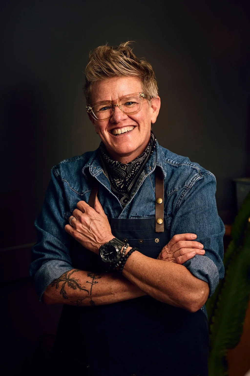 A smiling woman with short, styled hair wearing glasses, a denim shirt, and an apron, standing with arms crossed in front of a dark background.