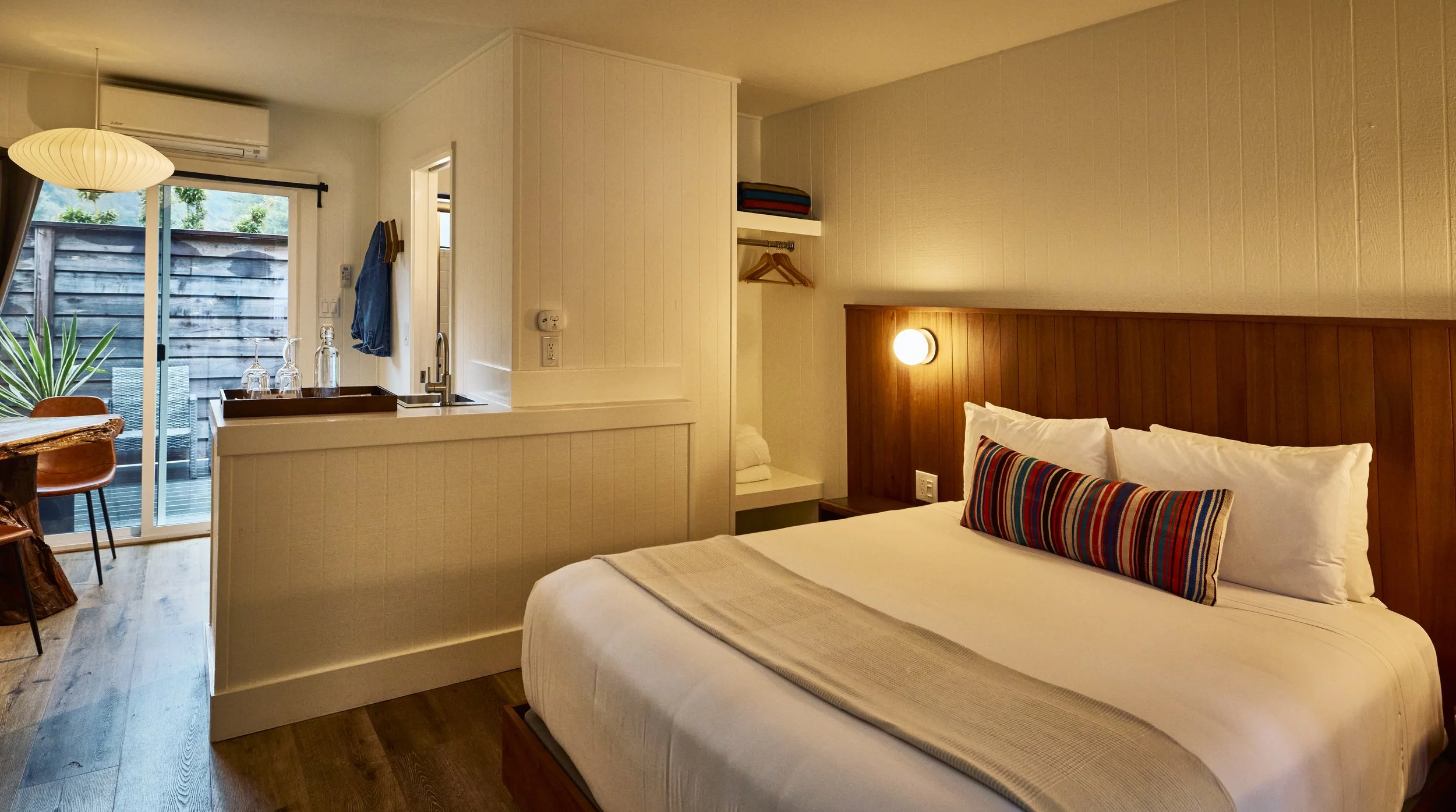 A cozy hotel room with a bed, pillows, and a wooden headboard. There is a wall-mounted light next to the bed, and a small nightstand with a shelf. The room has a sliding door leading to a small outdoor balcony with a table and chairs. The room features light-colored walls and wooden flooring.