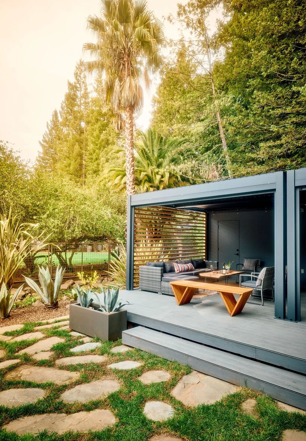 Outdoor patio area featuring a modern open-air structure with dark gray metal framing, a wooden slat privacy screen, and outdoor furniture including black wicker sofas, a wooden table, and armchairs. Surrounding the patio are lush green plants, agave in a gray planter, and a garden with stepping stones leading to the patio. Tall trees, including palms, and dense greenery backdrop the scene.