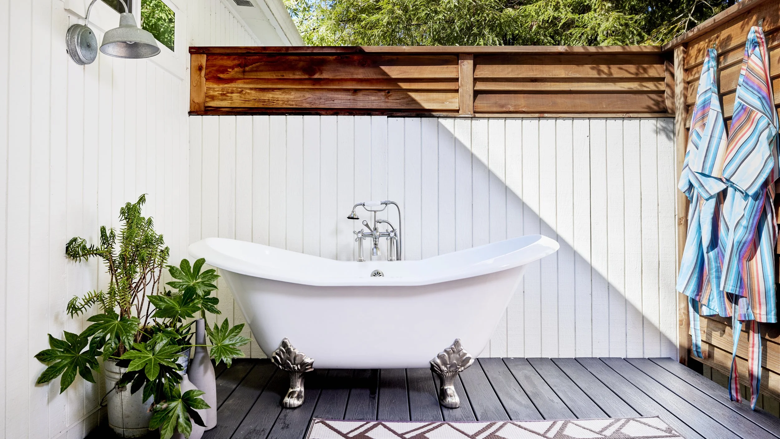 A vintage white clawfoot bathtub with clawfoot legs, placed on a dark wooden deck, with green potted plants beside it, set against a white paneled wall with a window and an outdoor shower fixture, and a wooden privacy fence with colorful striped towels hanging on it.