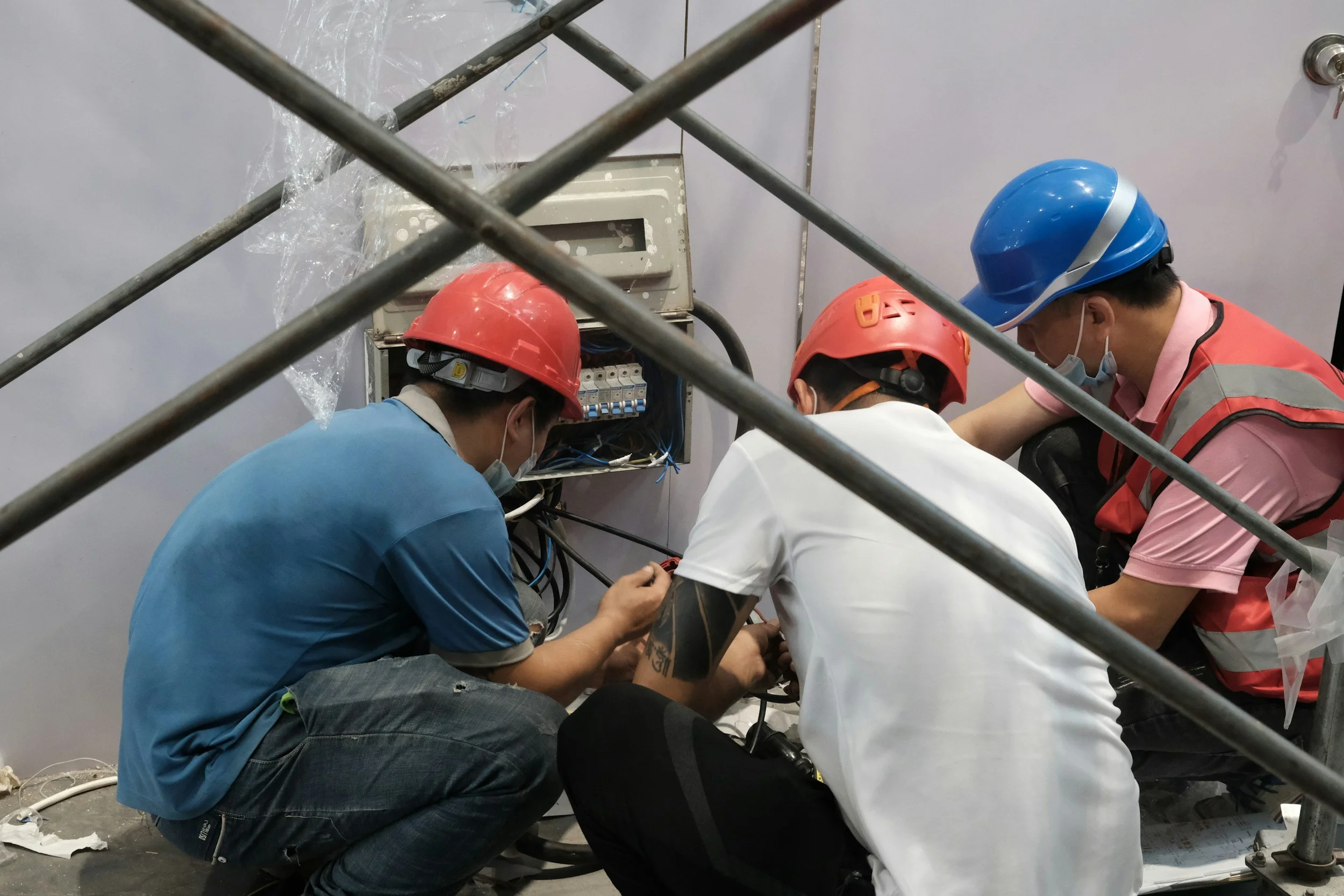 Four workers wearing hard hats and face masks are fixing electrical wiring inside an electrical panel.