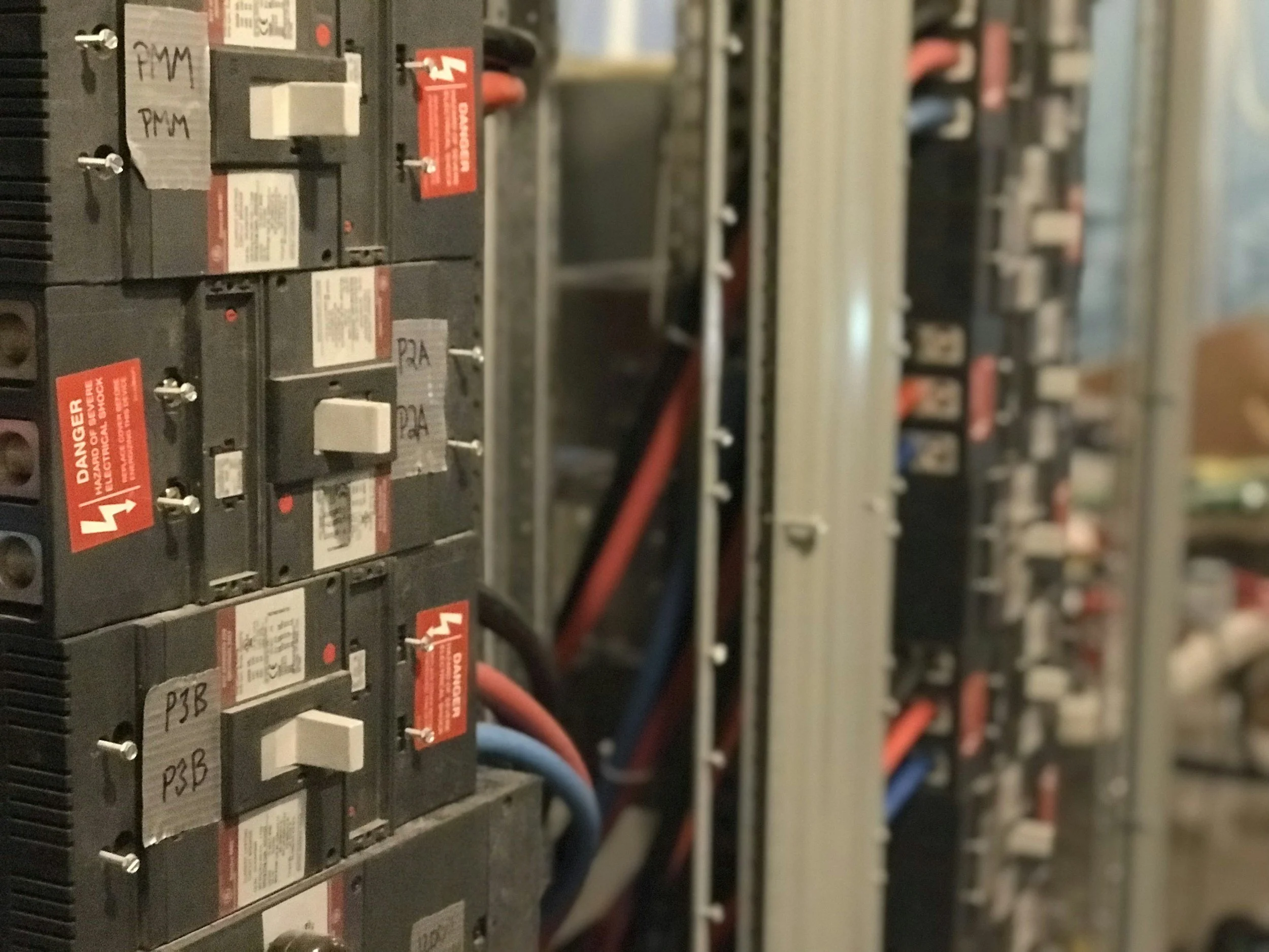 Close-up of a row of black electrical circuit breakers with white labels and red warning labels, connected with red and blue wires in an electrical panel
