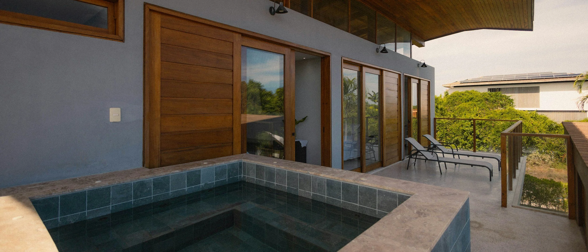 Balcony with two lounge chairs overlooking trees, with glass sliding doors and wooden accents, and a hot tub on the corner.
