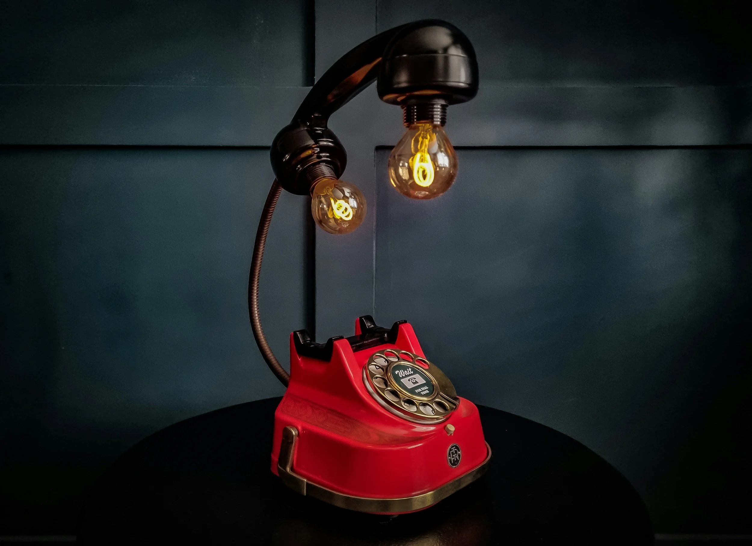 A vintage red rotary phone with a black dial, placed on a dark surface with a dark background. The phone's cord is visible, and the rotary dial features numbers and letters.