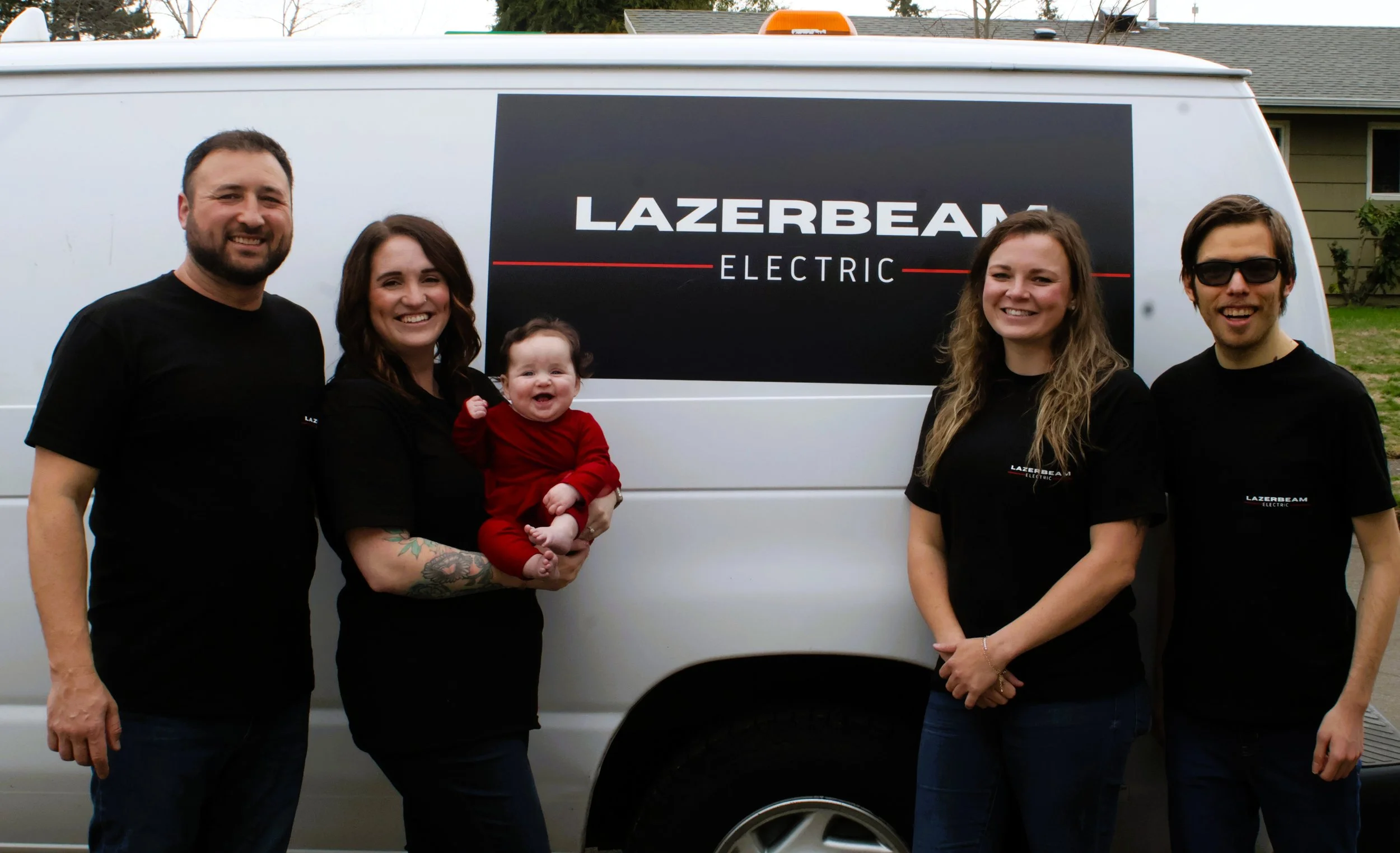 Group of four adults and one baby standing in front of a white van with a 'Lazerbeam Electric' sign on it. The adults are smiling, and the baby is wearing a red outfit.