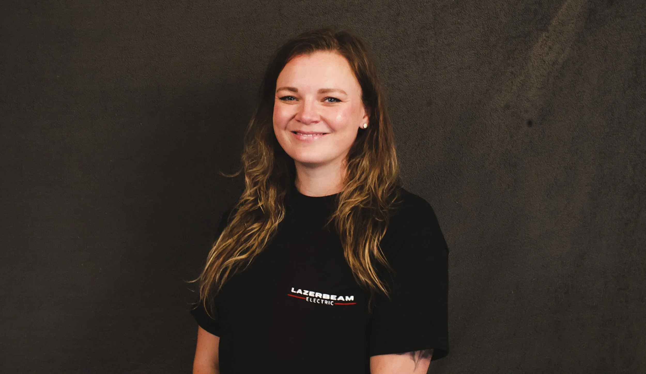 Smiling woman with wavy brown hair wearing a black T-shirt with 'LAZERBEAM ELECTRIC' logo, standing against a dark wall.