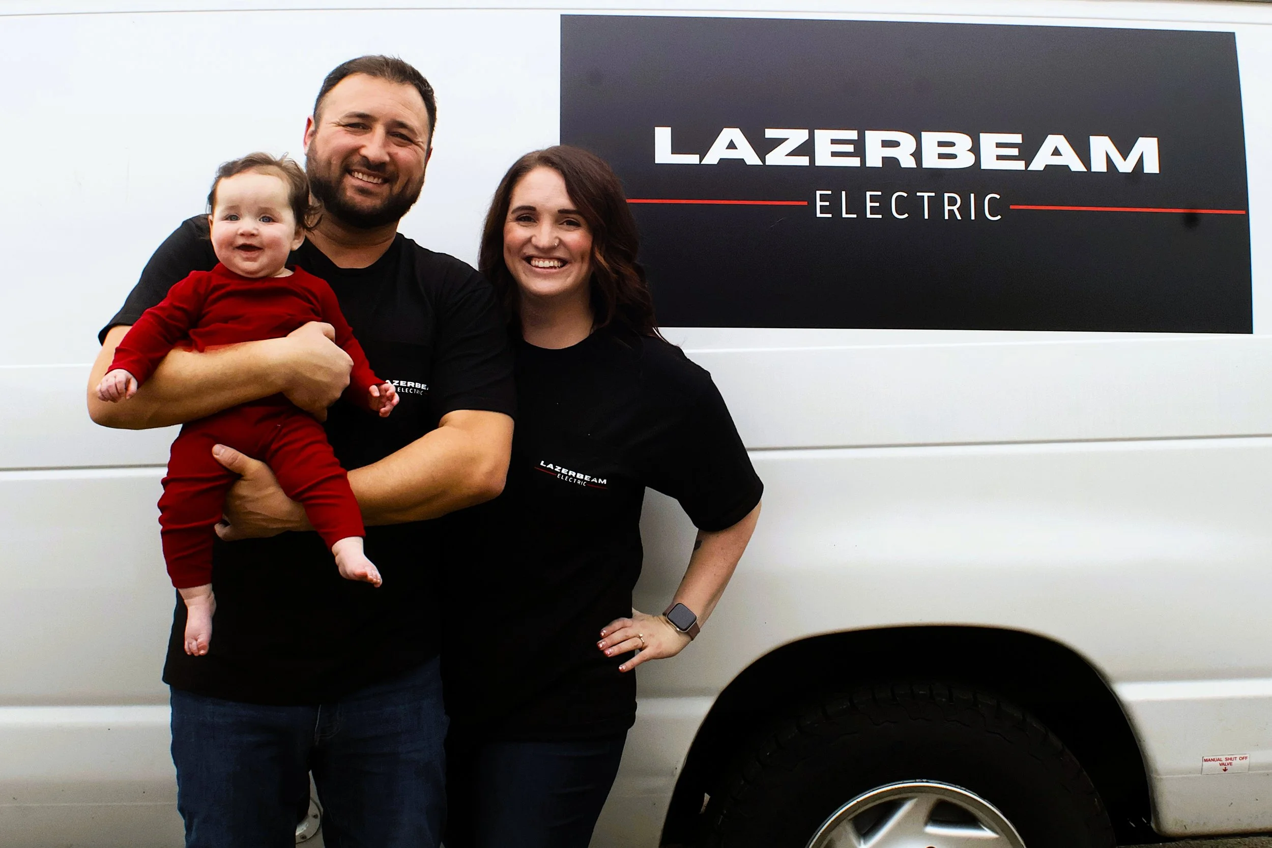 Family of three posing in front of a Lazerman Electric vehicle, the man is holding a baby girl, and the woman stands beside them with her hand on her hip.