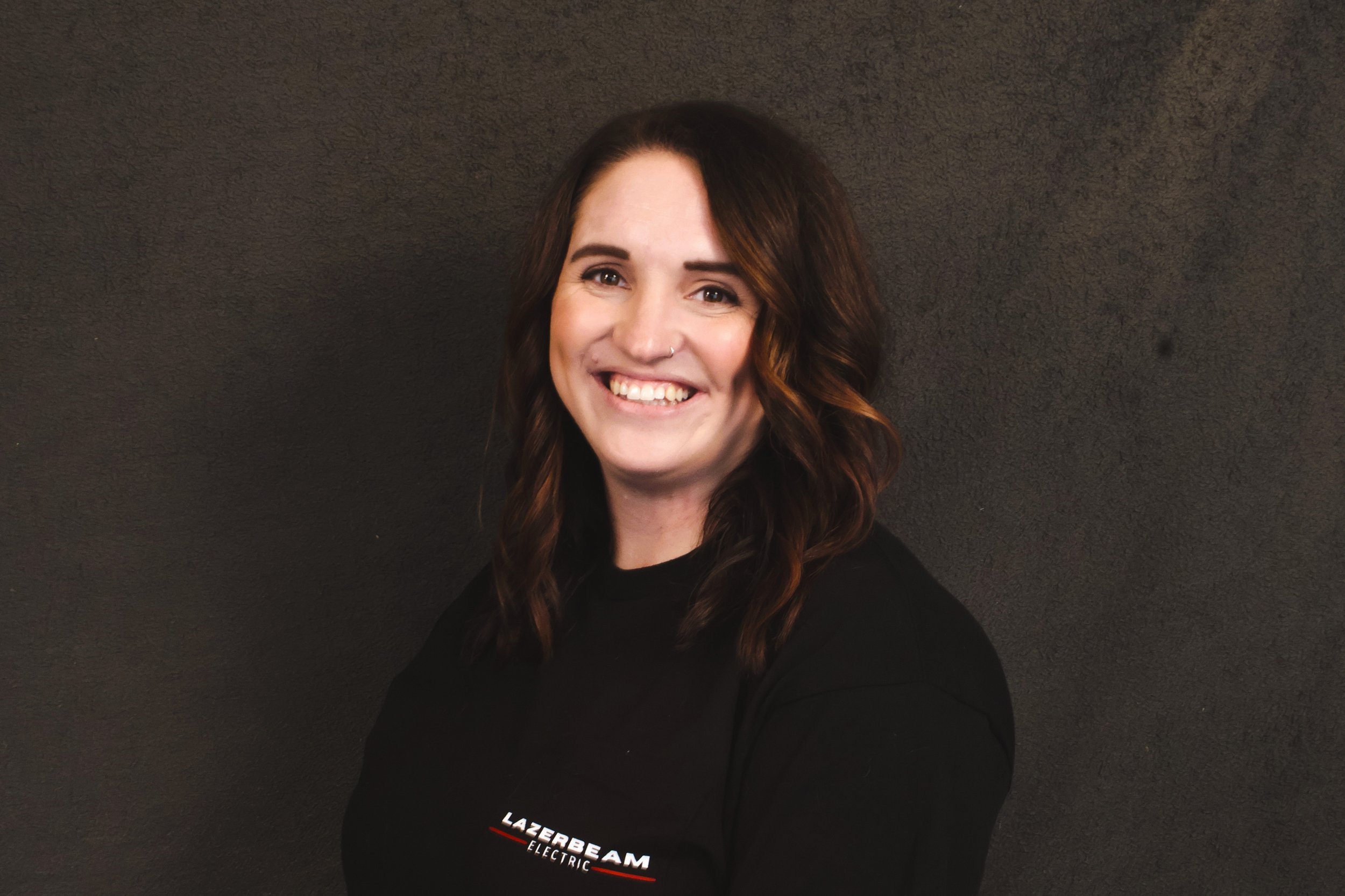 A smiling woman with wavy brown hair, wearing a black shirt with the words "Lazerbeam Electric" on it, standing against a dark wall.