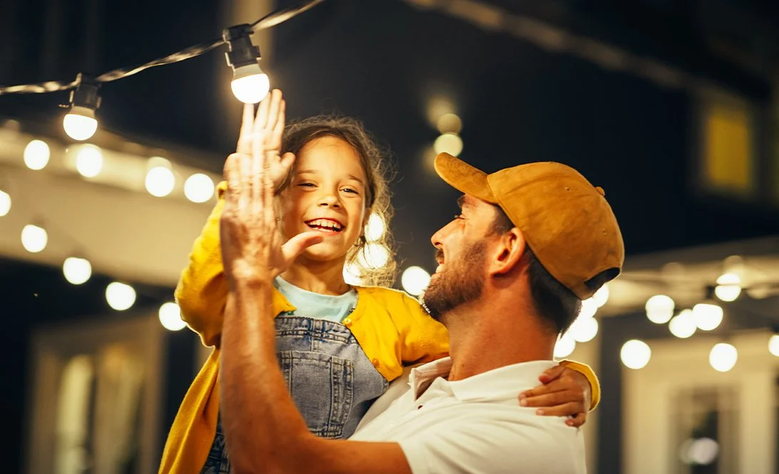A man holding a smiling young girl high in the air during an evening outdoor gathering with string lights.