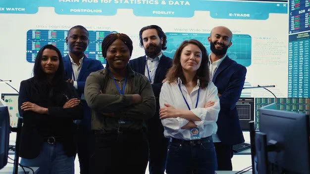 Group of five diverse professionals standing in front of a data and statistics display board at a control center or office.