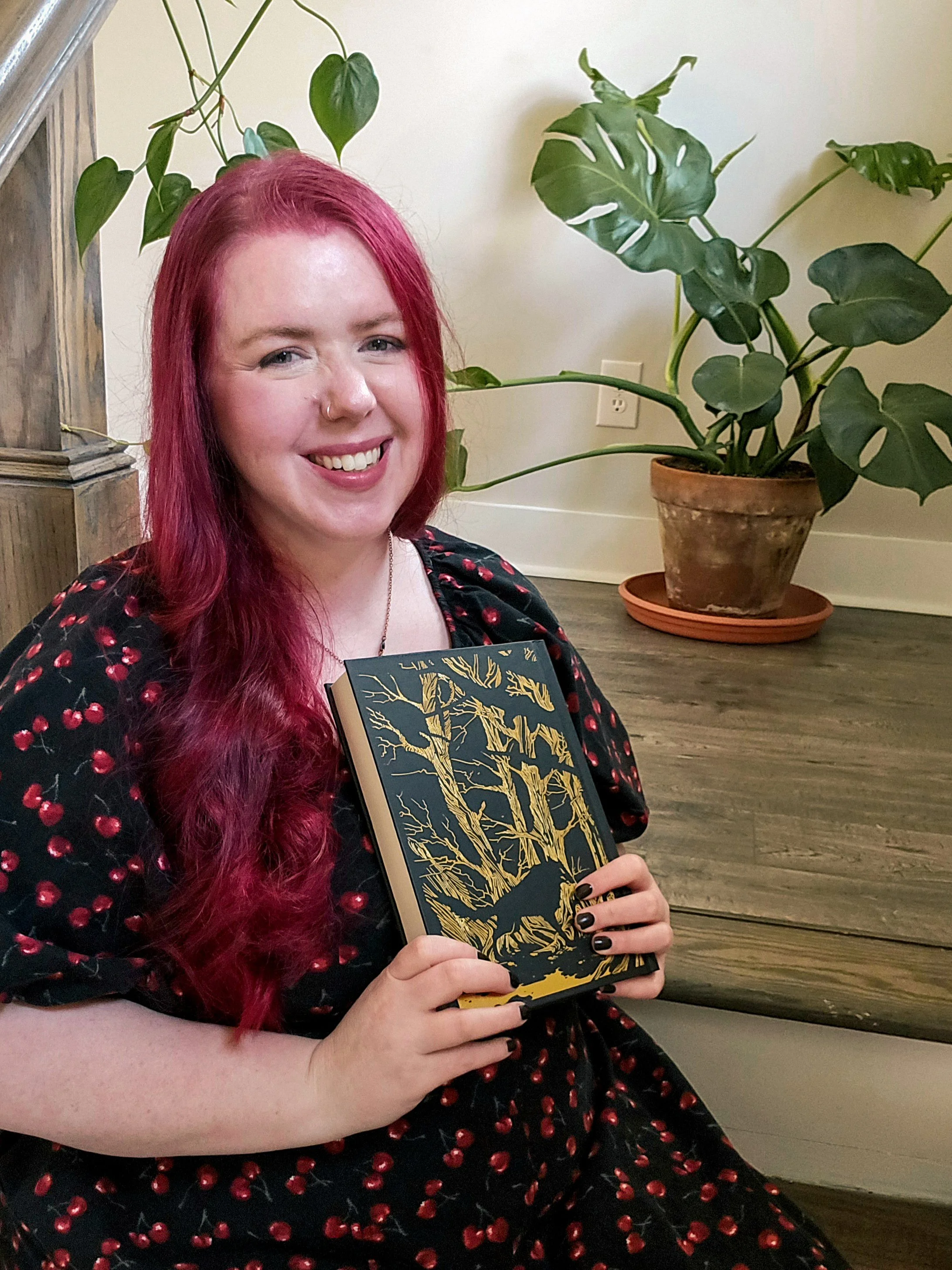 A woman with long red hair and a nose piercing smiling, holding a book with a gold tree design on its cover, sitting indoors near a large potted plant with dark green leaves.