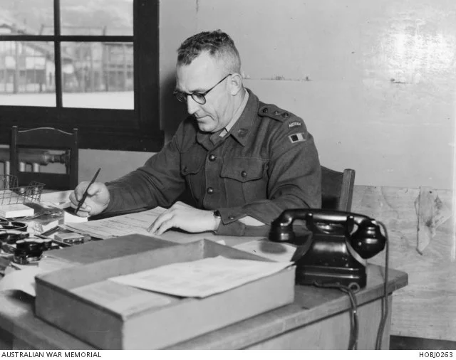 An unidentified Australian Captain (Capt) of the British Commonwealth Occupation Force (BCOF), concentrating on paperwork at his office desk. Note he is wearing a colour patch on his left shoulder that indicates he is probably a member of the Royal C