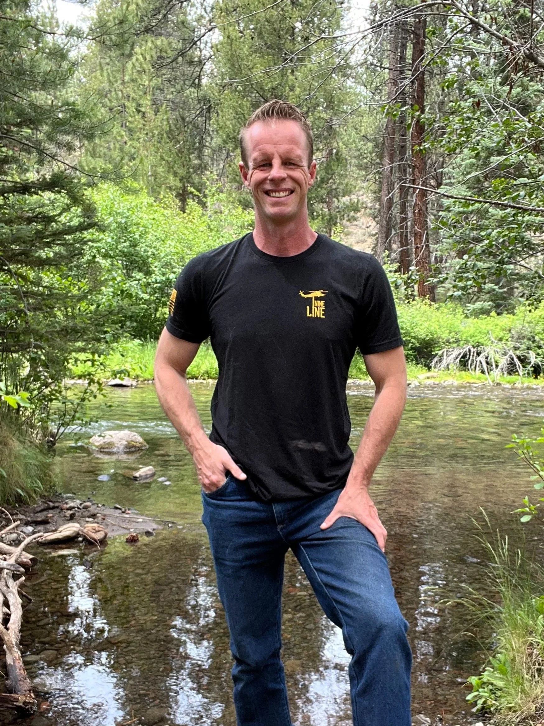 A man standing in a shallow stream surrounded by green trees and foliage, smiling at the camera, wearing a black t-shirt and blue jeans.
