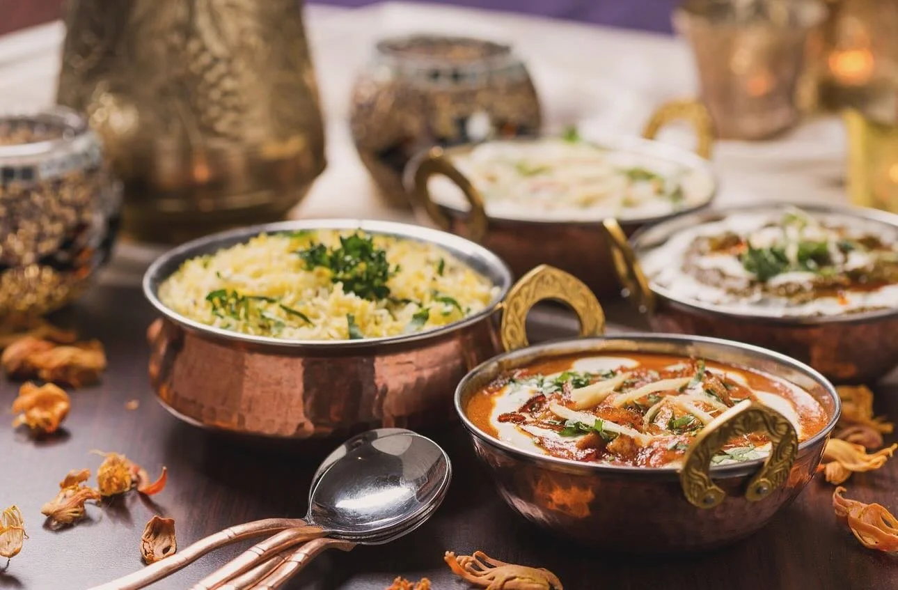 Several traditional Indian copper bowls filled with various curries and rice dishes on a dark table, with spoons and scattered flower petals.
