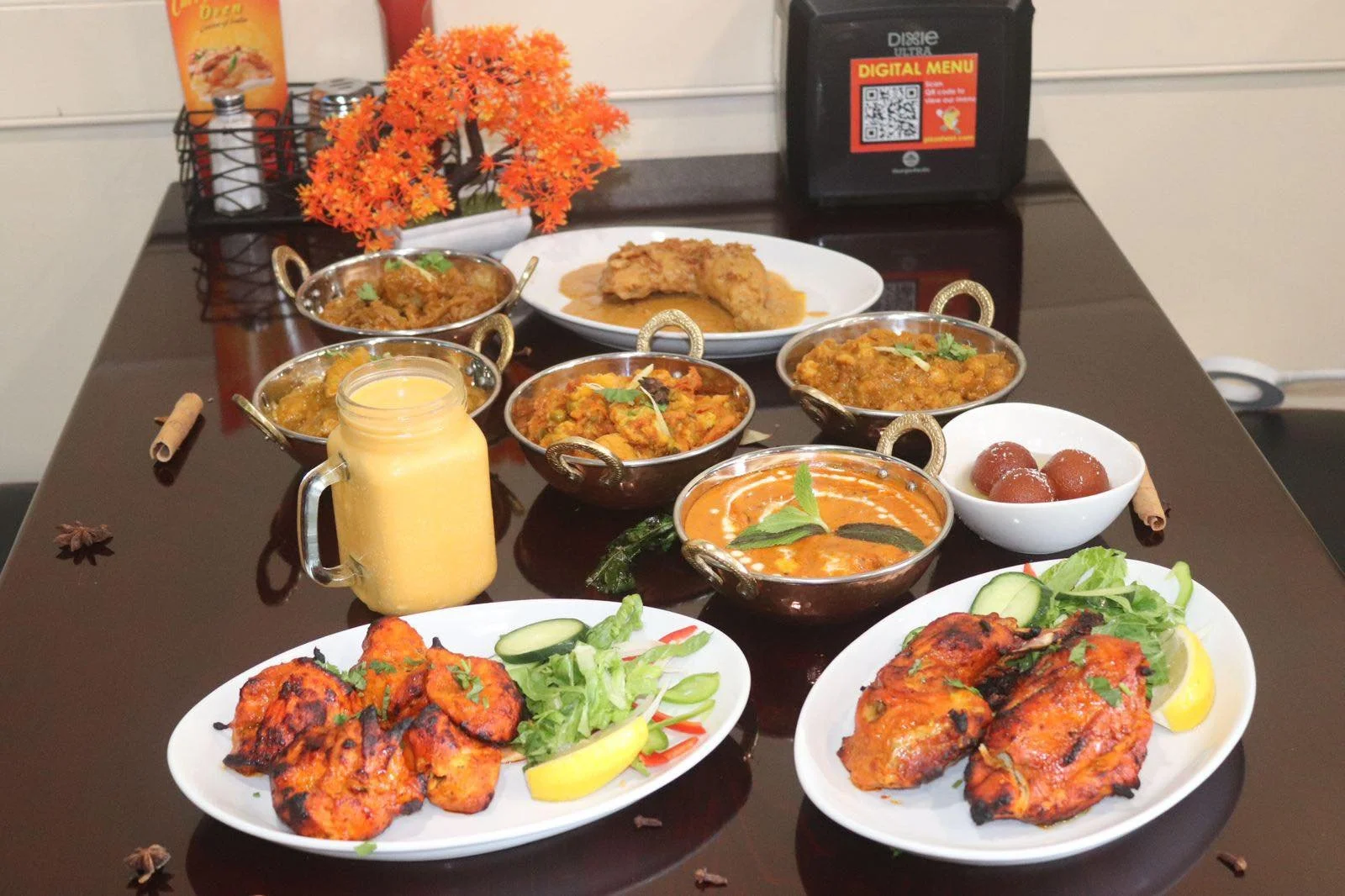 Indian meal spread on a dining table including two plates of tandoori chicken, various curries in metal bowls, a jar of mango lassi, and side dishes like cucumber slices, lettuce, lemon wedges, a bowl of gulab jamun, and a plate of fried chicken with