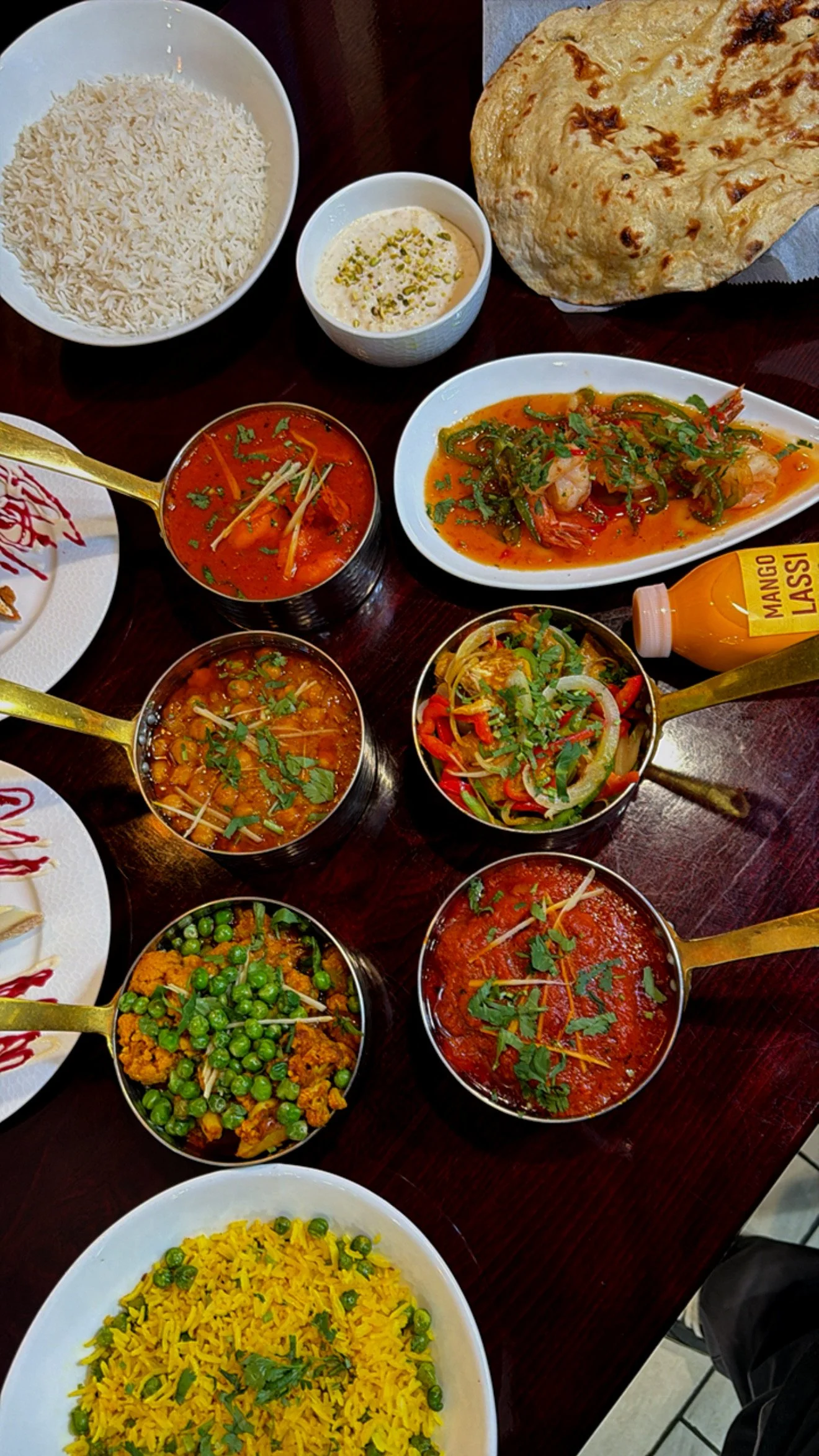 A variety of Indian dishes including white rice, yellow rice, naan bread, dum aloo curry, mixed vegetable curry, spiced peas and cauliflower, vegetable salad, various meat curries, and a bottle of mango lassi on a dark wooden table.