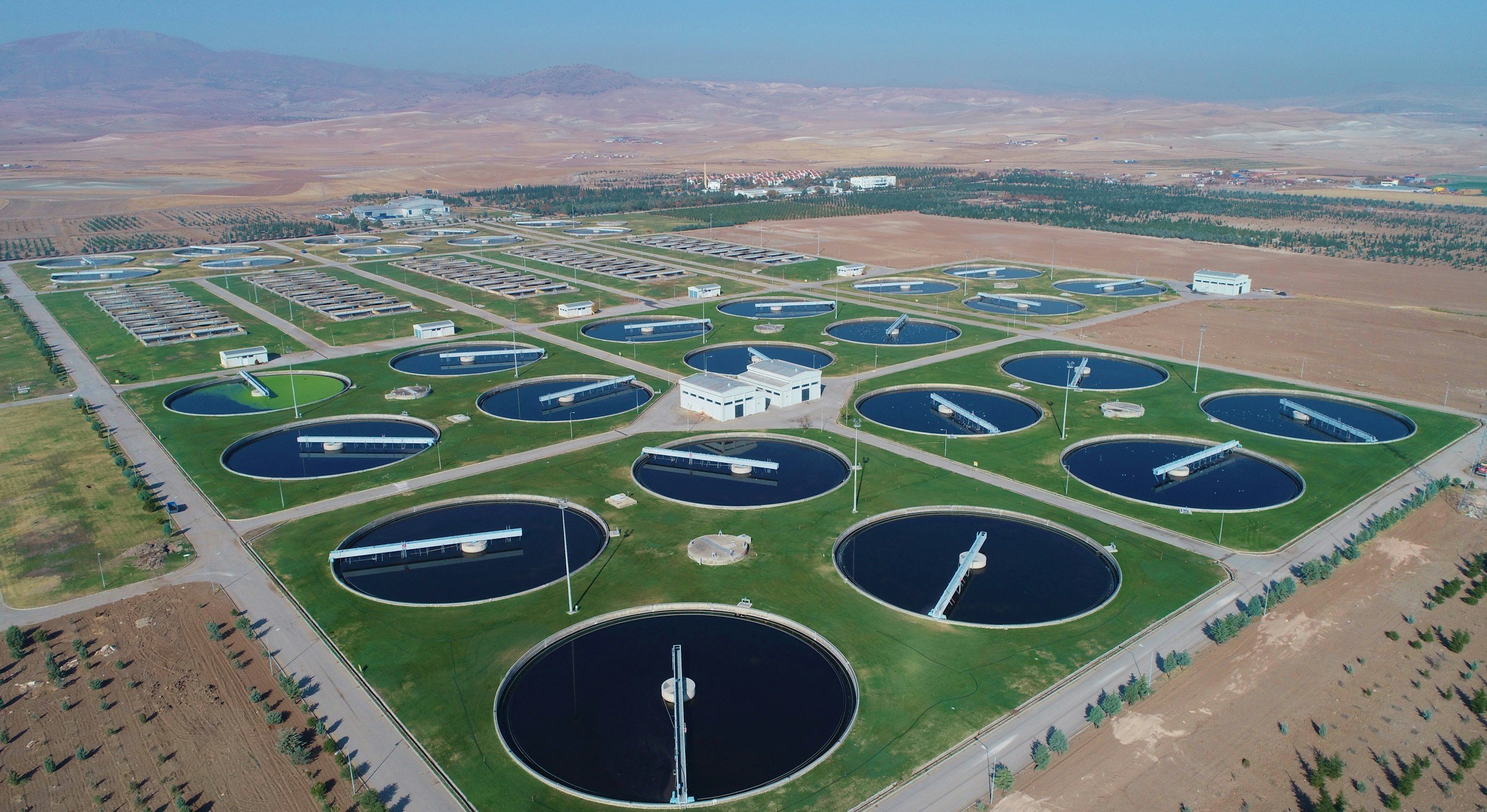 Aerial view of a large wastewater treatment plant with circular treatment tanks, surrounded by roads, greenery, and barren land.