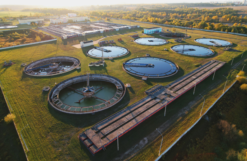 Aerial view of large circular water treatment tanks at a wastewater treatment plant during daytime, with surrounding greenery and industrial buildings in the background.