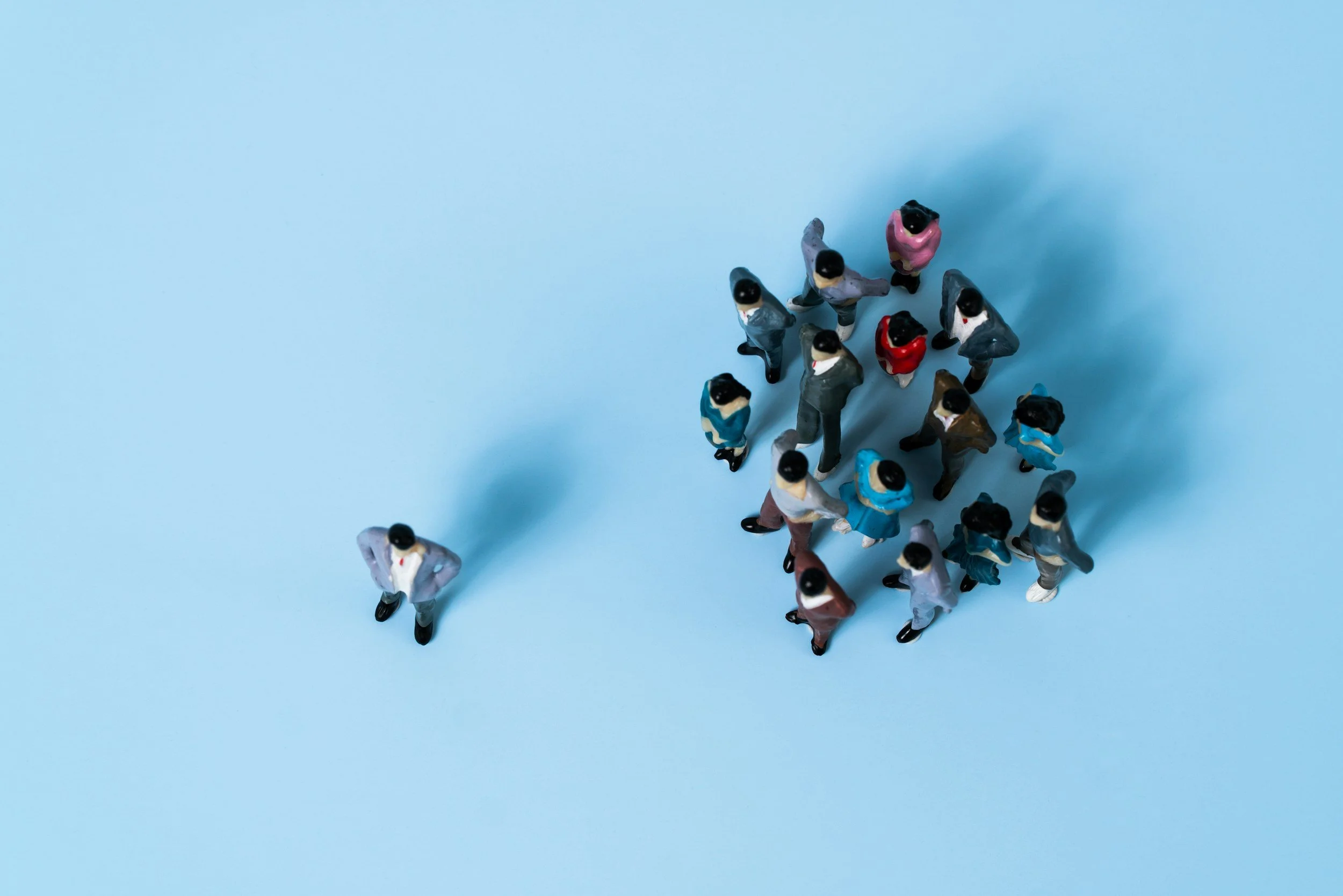Top-down view of a group of miniature figurines of people in business attire on a light blue background, arranged in a loose cluster.