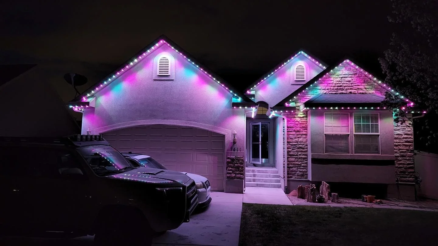 Nighttime photo of a house decorated with multicolored string lights along the roofline, with a garage door, a front porch with steps, and a few parked cars in the driveway.