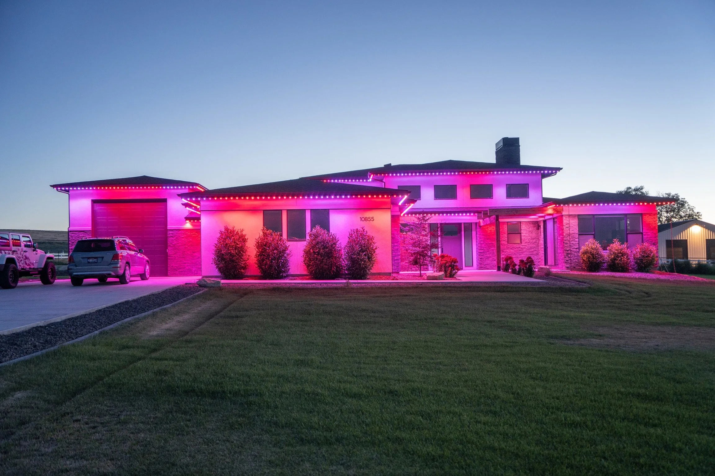 A modern house decorated with pink and purple neon lights at dusk. The house features a garage, multiple windows, and a well-manicured lawn.