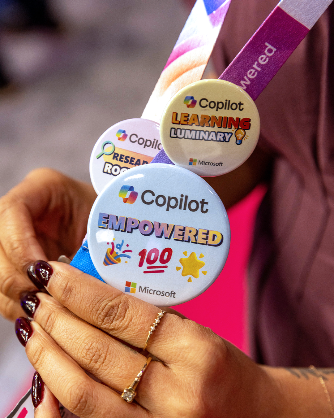 Close-up of a person's hand holding three motivational buttons, each featuring the Microsoft and Copilot logos and positive messages about learning, research, and empowerment.