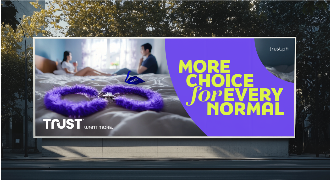 A billboard advertisement featuring two women sitting on a bed and talking, with purple fuzzy handcuffs in the foreground and the text 'More choice for every normal' in large yellow and green letters. The brand 'Trust' and the website 'trust.ph' are also displayed.