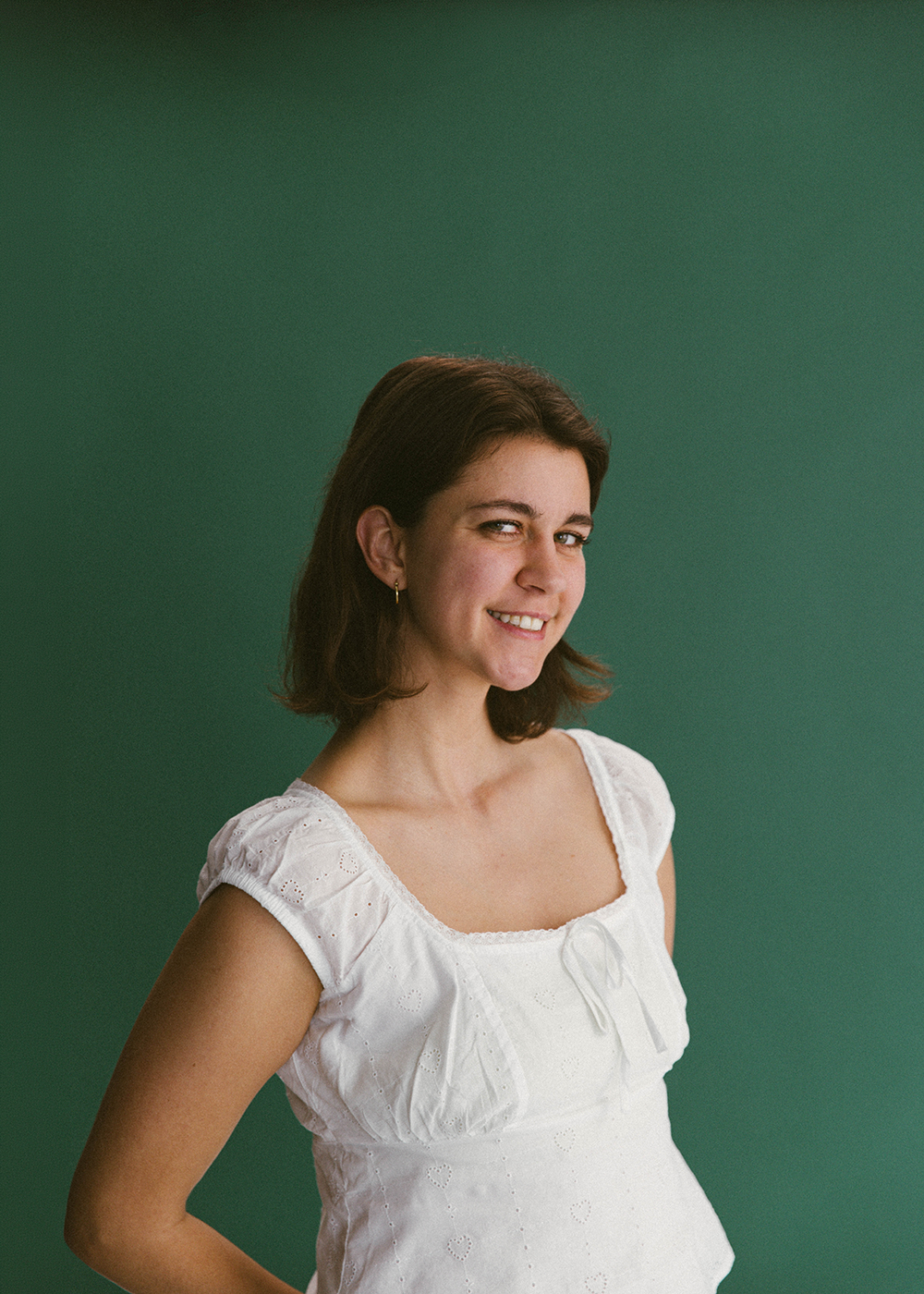 Smiling young woman with shoulder-length dark brown hair, wearing a white eyelet dress with puffed sleeves, standing against a solid green background.