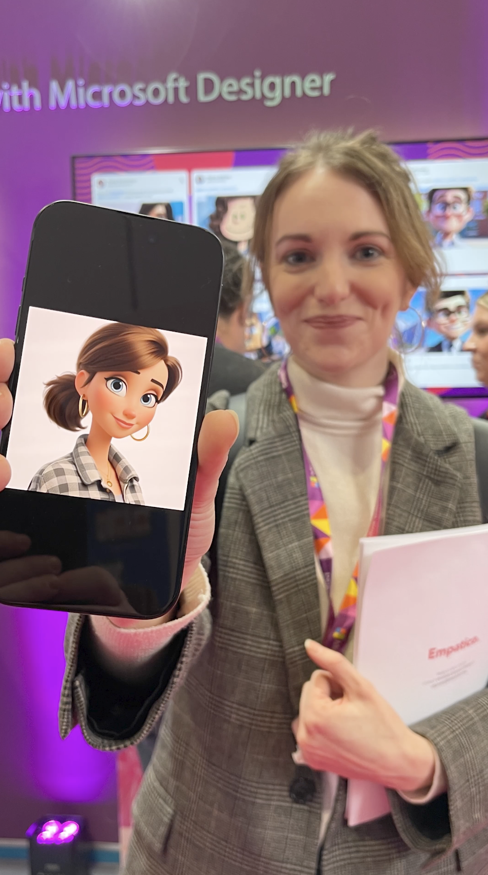 A woman in a blazer smiling and holding a folder in front of her, displaying a phone with an animated character on the screen, at a Microsoft Designer event.