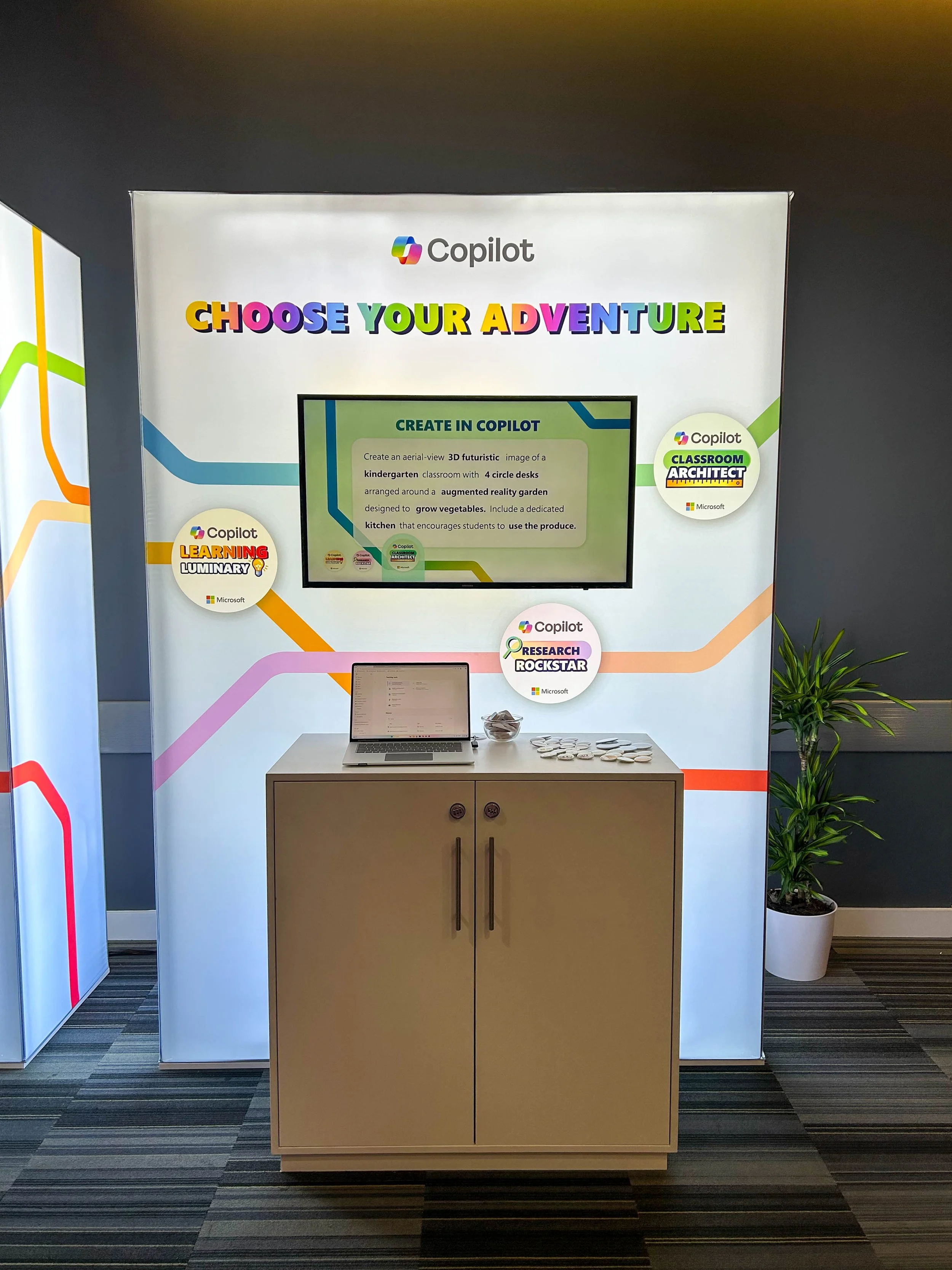Exhibit booth with a sign reading "Choose Your Adventure" at the top, featuring a screen with a promotional message about creating an aerial view 3D futuristic image for a kindergarten classroom with desks and a garden, bright colors, and national green, yellow, pink, and purple accents. A laptop, a bowl of snacks, and a potted plant are on a small table in front of the booth.