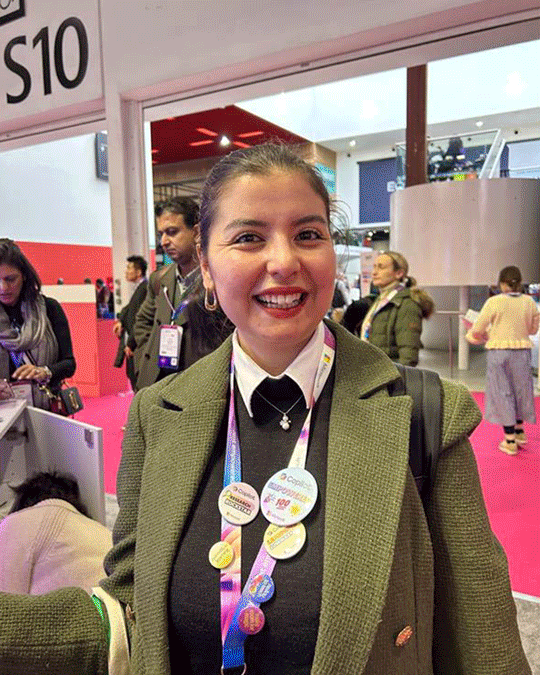 A smiling woman with a dark ponytail wearing a green blazer, white shirt, and black sweater at an indoor event with pink flooring, surrounded by people, some with badges or lanyards.