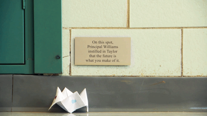 A paper origami boat on a classroom floor against a beige brick wall with a plaque that reads, "On this spot, Principal Williams instilled in Taylor that the future is what you make of it."