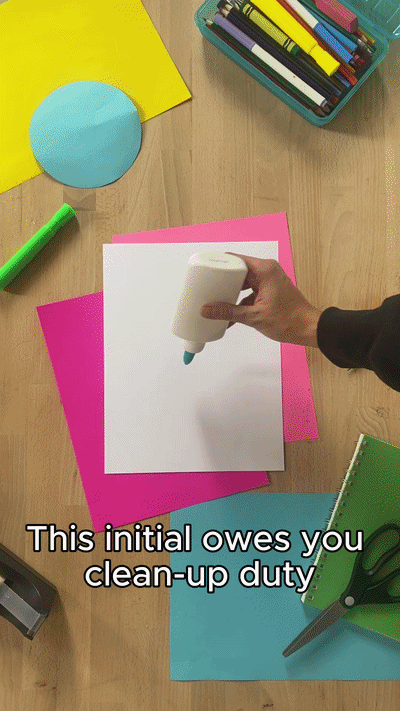 Hand holding a glue stick over a stack of colored papers on a wooden table surrounded by office supplies, including scissors, a green marker, and containers of markers and paper.