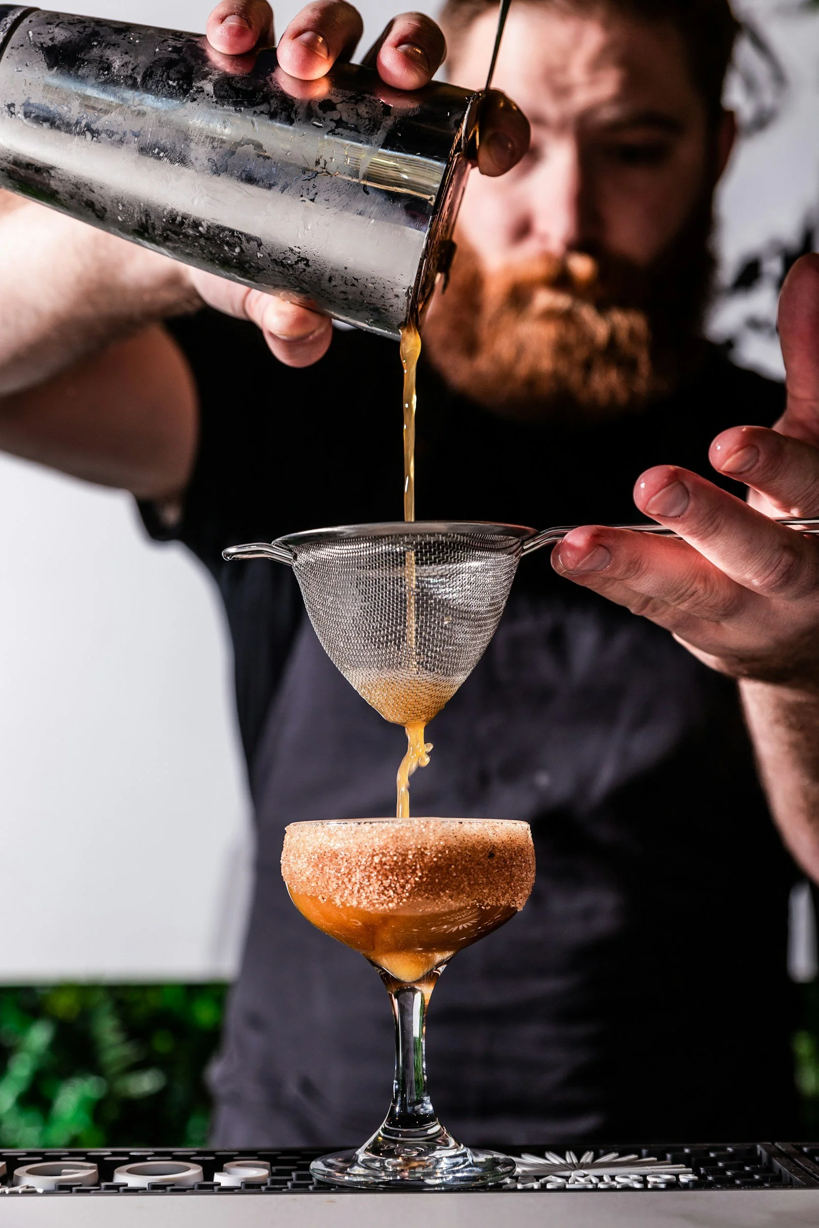 A bartender pours liquid through a fine strainer into a cocktail glass with salted rim, above a black bar mat with white lettering.